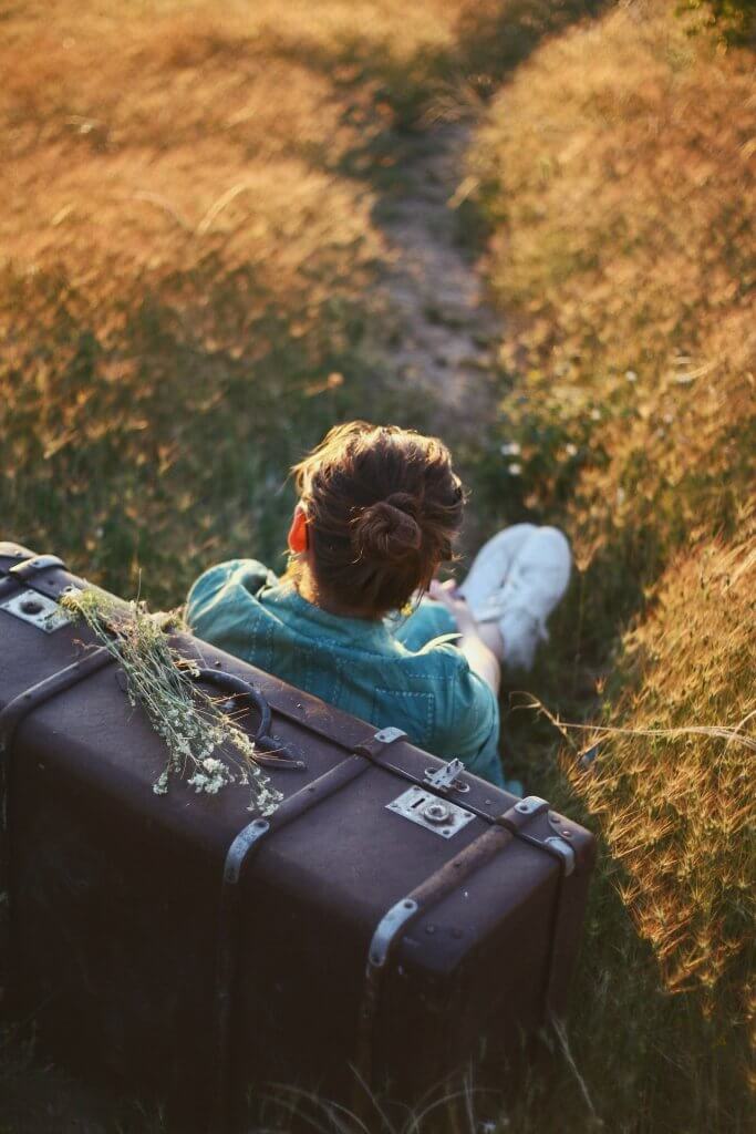 Femme assise adossée à sa valise sur un chemin de campagne