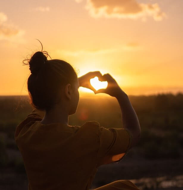 Jeune femme formant un coeur avec ses doigts face au soleil couchant